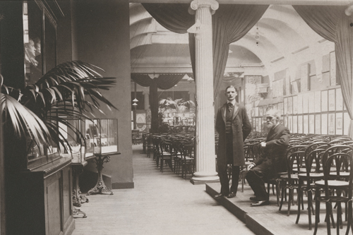 An old photograph of two men standing in an elegant, empty exhibition hall with rows of chairs and framed artworks on the walls.